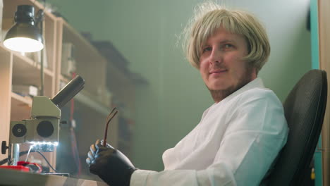 close-up view of a technician in a blonde wig and removing glasses, black gloves while seated at a lab desk with a microscope