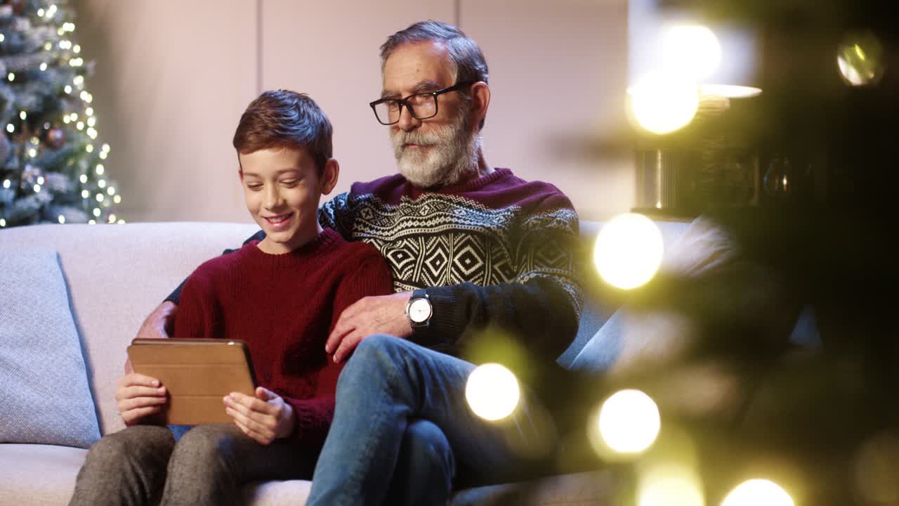 retrato del viejo abuelo con un alegre nieto adolescente sentado en una habitación decorada tocando en una tableta eligiendo regalos de navidad y navegando en internet cerca del brillante árbol de año nuevo