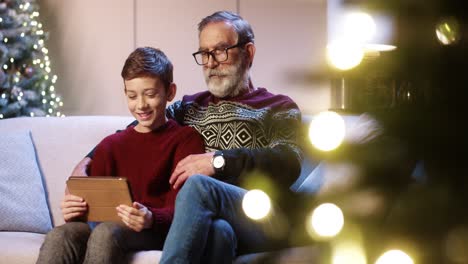 portrait of old grandpa with joyful teen grandson sitting in decorated room tapping on tablet choosing christmas presents and browsing on internet near glowing new year tree