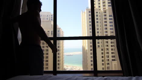 a morning light fills a hotel room as a man stands by the window, gazing out at towering buildings and a tranquil sea in the distance