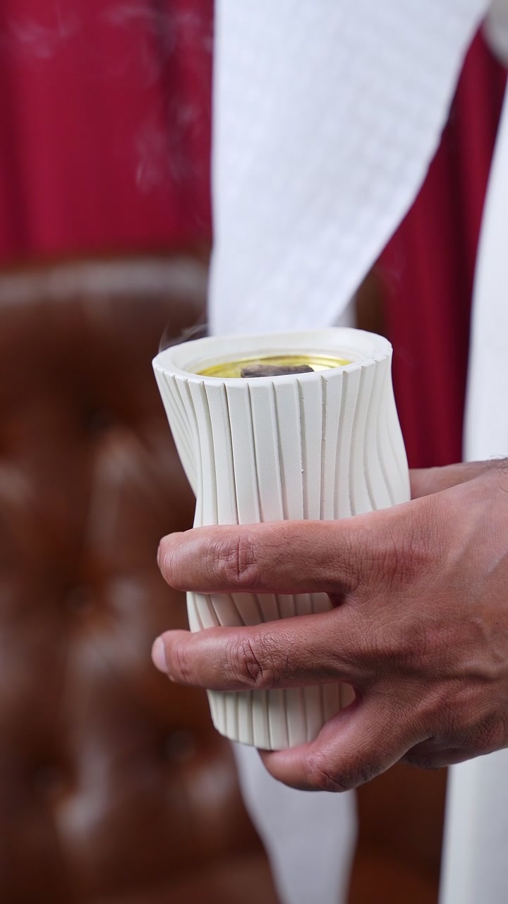 Close-up of an Arab man's hand holding a white textured incense burner. Fragrant smoke rises against traditional white garments in a modern indoor setting, highlighting Middle Eastern culture.