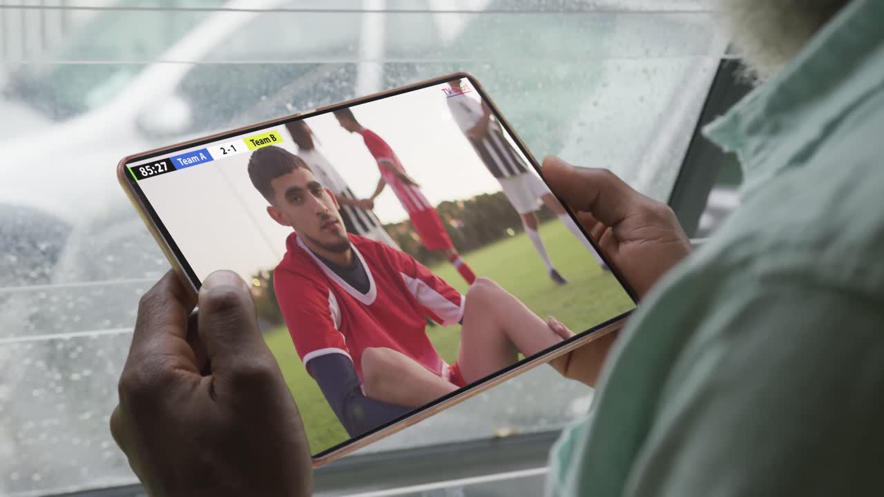 다양한 남성 축구 선수들과 함께 태블릿을 사용하는 아프리카계 미국인 남자가 화면에서 경기를 하고 있습니다.