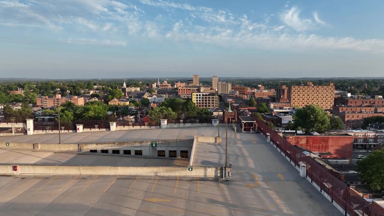 Ripresa aerea con drone in ascesa di un parcheggio multipiano con veicoli parcheggiati e skyline di una città americana sullo sfondo. Tramonto a Lancaster City, PA, USA. Campo lungo