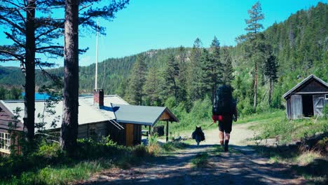 Backpacker-With-His-Dog-Passing-On-The-Road-Through-Rural-Village-Near-Forest-Mountain