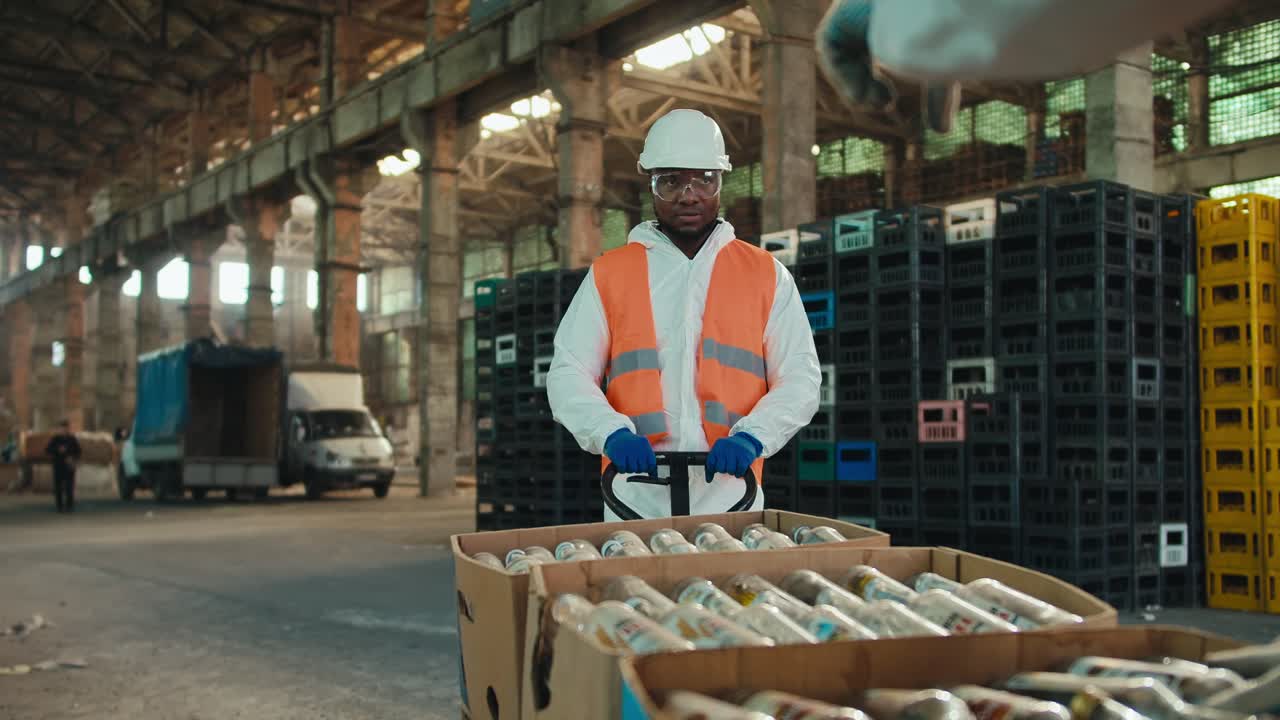 A man with Black skin in a white protective uniform and an orange vest carries boxes with sorted glass garbage and bottles on a special wheelbarrow at a waste processing and sorting plant
