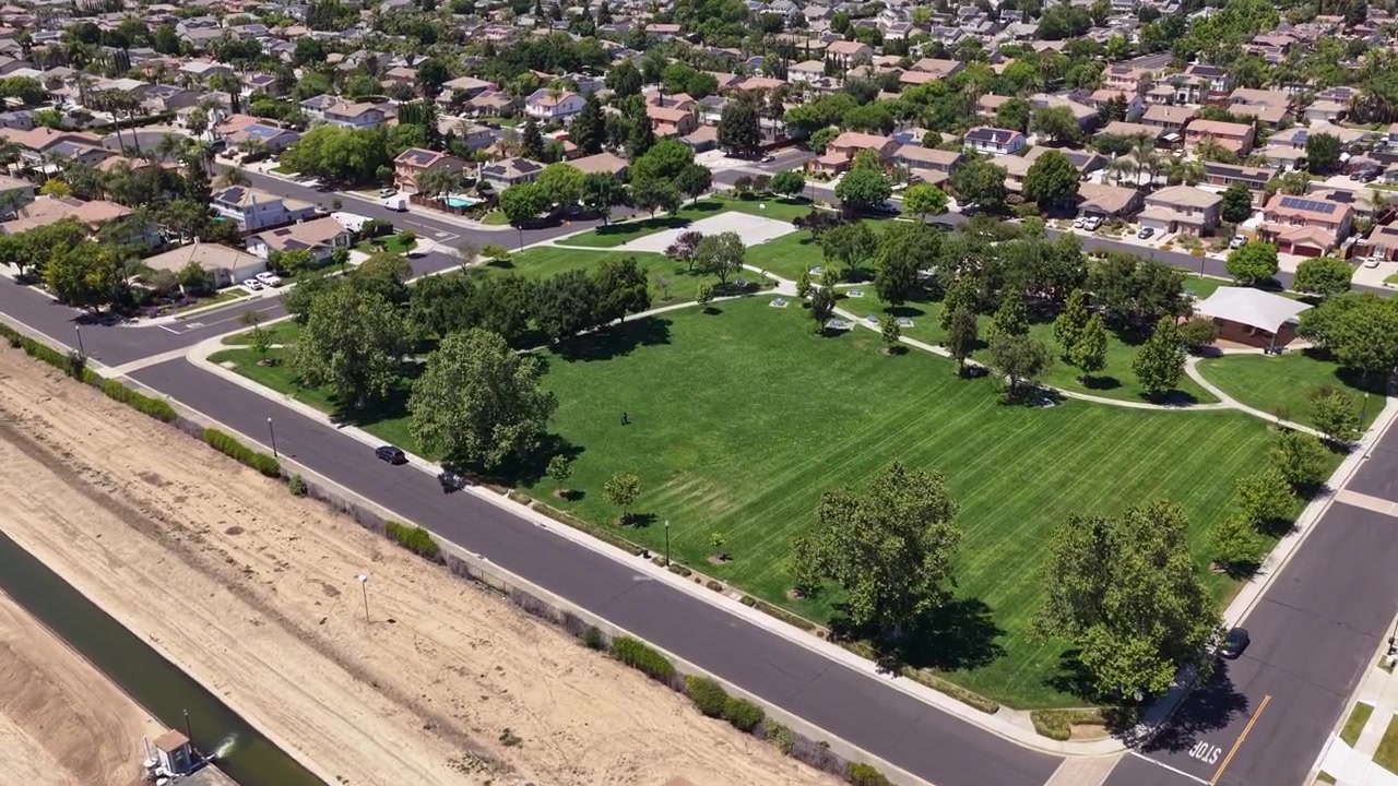Aerial drone descending view of a lush green city park surrounded by residential suburban houses and streets in Brentwood, California on a sunny day with an irrigation canal nearby