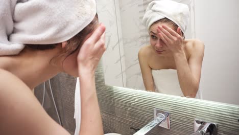 A-woman-standing-in-the-bathroom-near-the-sink-in-white-towel-on-head-and-body-washes-her-face-with-water-from-the-tap.-Looking-to-the-mirror-reflection-and-smiling