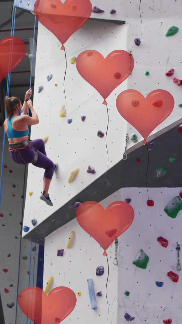 Palloncini rossi a forma di cuore che fluttuano sullo sfondo di una donna caucasica in forma che fa arrampicata in palestra.