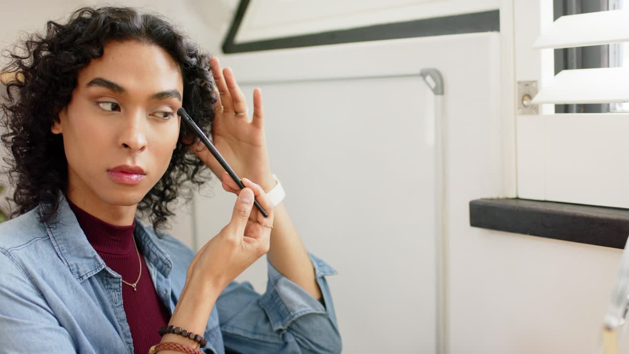 Non-binary person reaching for eyeshadow brush dipping into palette, blending into crease at vanity