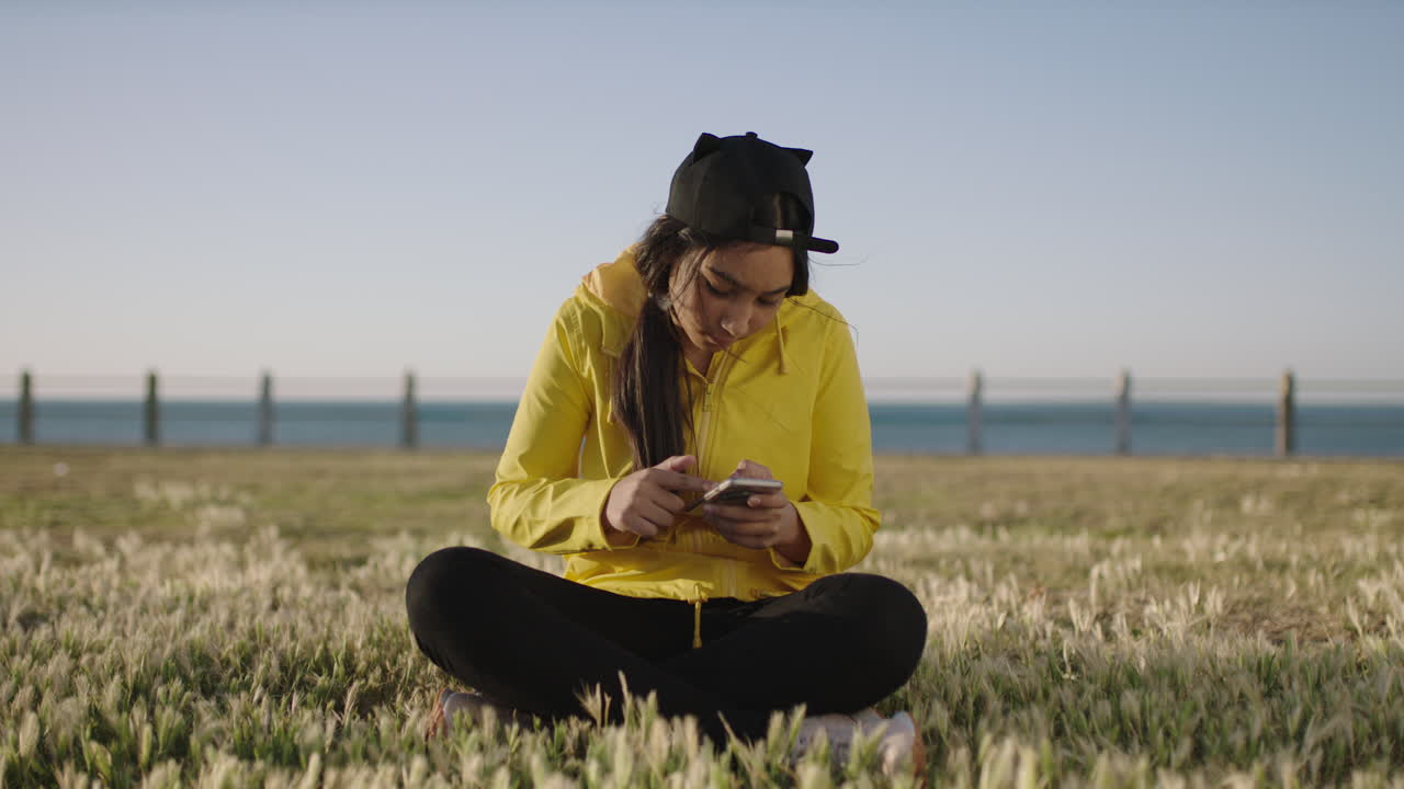 portrait of mixed race teenage girl sitting at seaside park texting browsing using smarphone social media app enjoying relaxed