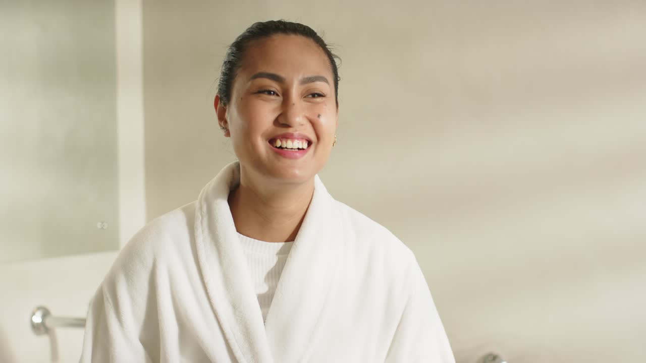 Smiling Asian woman wearing bathrobe raising arm smoothing hair twisting ponytail into bun at home