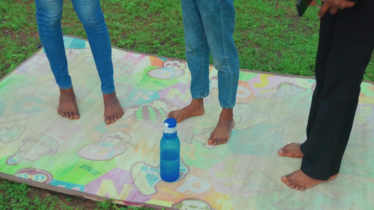 Barefoot mother child with adult guide on mat closeup feet around water bottle target, three participants in jeans and trousers on grass, coach directing playful steps, counting and timing, energetic