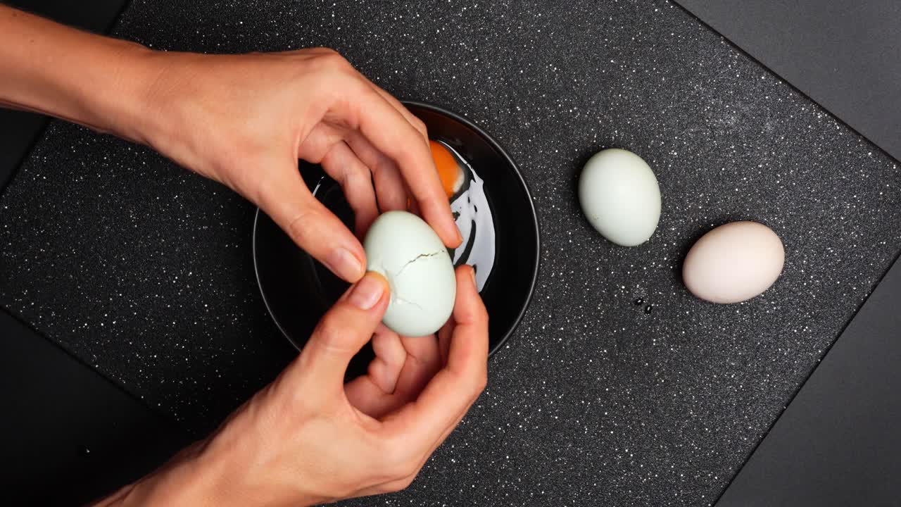 Cracking egg into bowl overhead. Hands cracking fresh egg into black bowl on dark surface