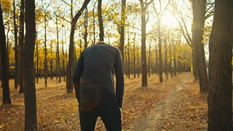 from behind a happy man with curly hair in black sports autumn clothes in an autumn forest with fallen leaves rests after his jog and prepares for new sports activities on a sunny day in autumn