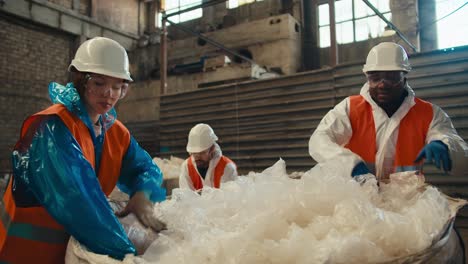 A-brunette-girl-in-a-blue-uniform-and-a-white-protective-helmet-together-with-her-male-colleagues-in-orange-vests-sorts-and-draws-cellophane-garbage-at-a-waste-processing-and-sorting-plant