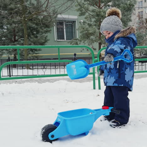 caucasian toddler in warm winter suit stands outdoors. lovely male kid picks up snow with his shovel.