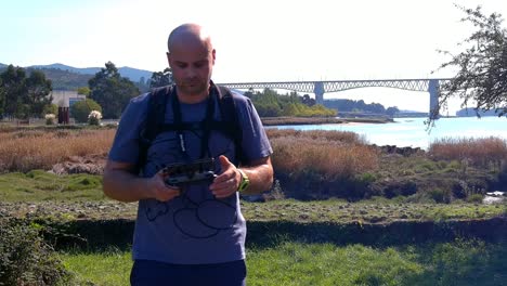 Piloto-De-Drones-Masculino-De-40-Años-Haciendo-Ajustes-E-Inspeccionando-Mirando-Directamente-A-La-Cámara-Con-Un-Puente-Y-El-Río-Detrás,-Soleada-Tarde-De-Verano,-Toma-Frontal