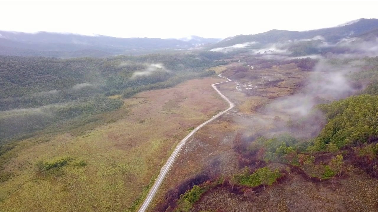 Volo aereo su montagne nebbiose e foresta verde e rossa in Tasmania, Australia, ripresa a lunghissima distanza in avanti e in picchiata