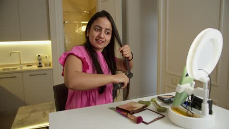 A-happy-brunette-Indian-girl-in-a-pink-dress-sits-at-a-table-and-smoothes-her-hair-with-a-special-straightener-during-her-video-blog-about-cosmetics-and-beauty-during-the-day-in-a-modern-apartment