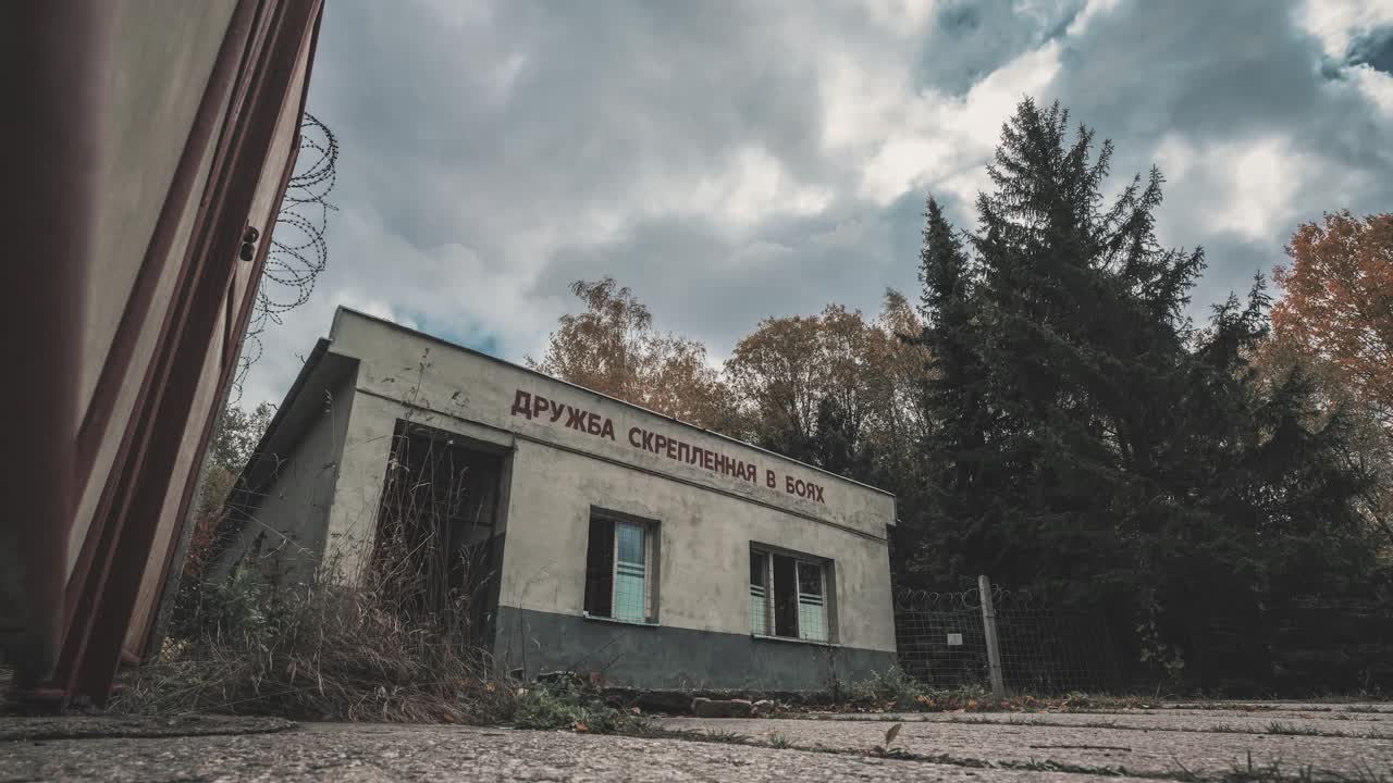 Timelapse of an abandoned, soviet-era military building with Russian text, set against a backdrop of overgrown foliage and tall pine trees under a cloudy sky.
