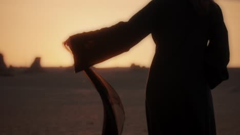 a girl holding a black scarf in her hand walking barefoot in the desert wearing an abaya while sunset stunning lens flare medium shot fossil dunes