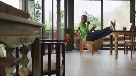 african american senior man sitting at dining table, relaxing with feet up, using smartphone