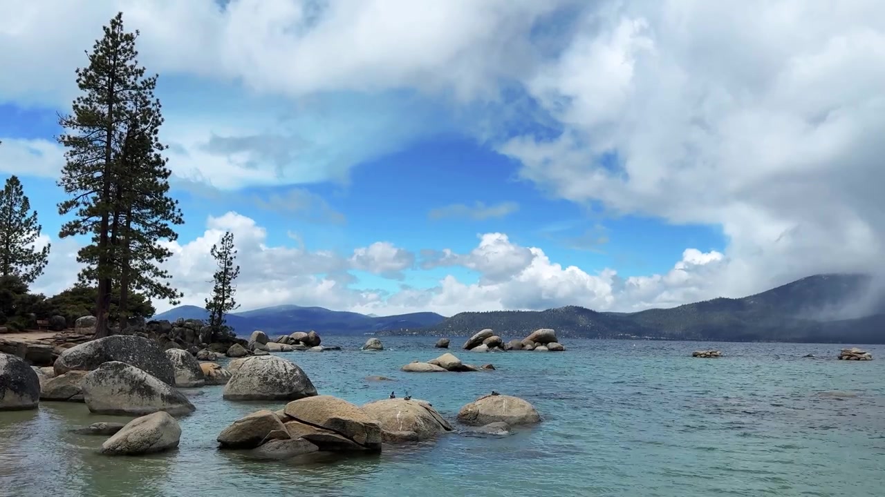 Batu-batu granit besar terhampar di air pirus sebening kristal di Pantai Sand Harbor, Danau Tahoe, dengan latar belakang pegunungan berhutan dan awan yang dramatis.