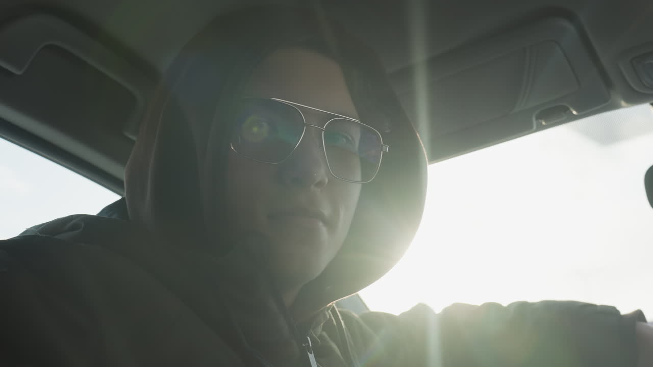student wearing hoodie and tinted glasses turns head back confidently over shoulder while gripping steering wheel inside sunlit car cabin overlooking snowy urban background