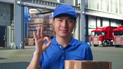 close up of asian male courier in blue uniform showing okay gesture and smiling while delivering a carton, outside of logistics distributions warehouse