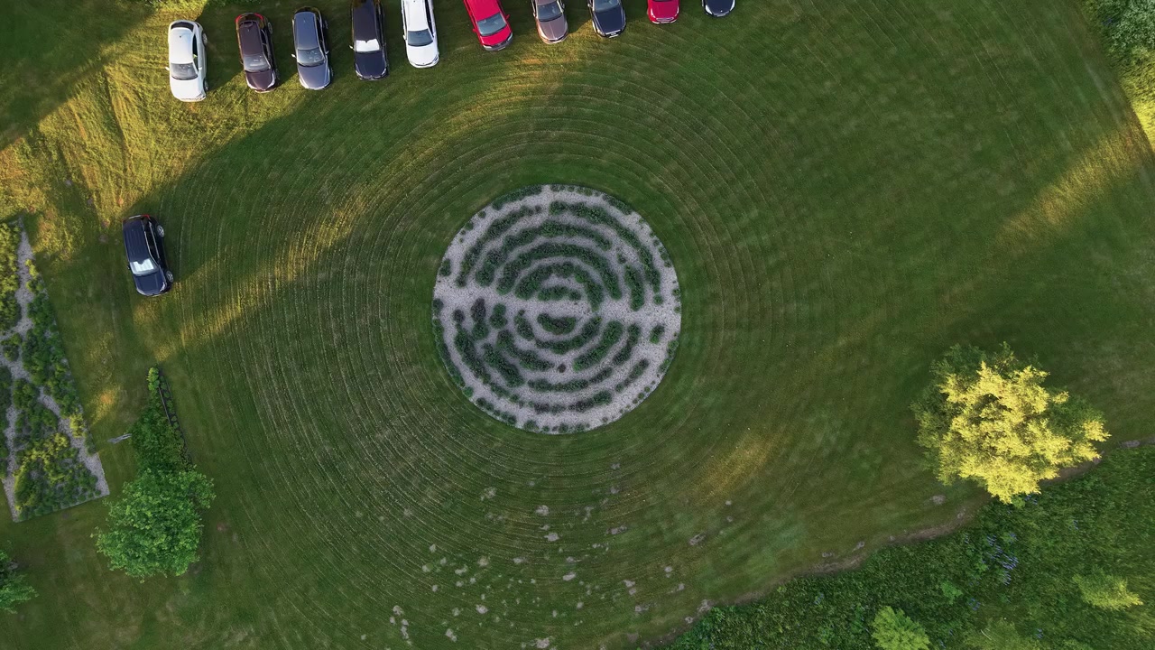Drone video captures a symmetrical labyrinth garden surrounded by manicured grass and several parked cars, filmed in soft late afternoon sunlight with crisp, vibrant colors from above.
