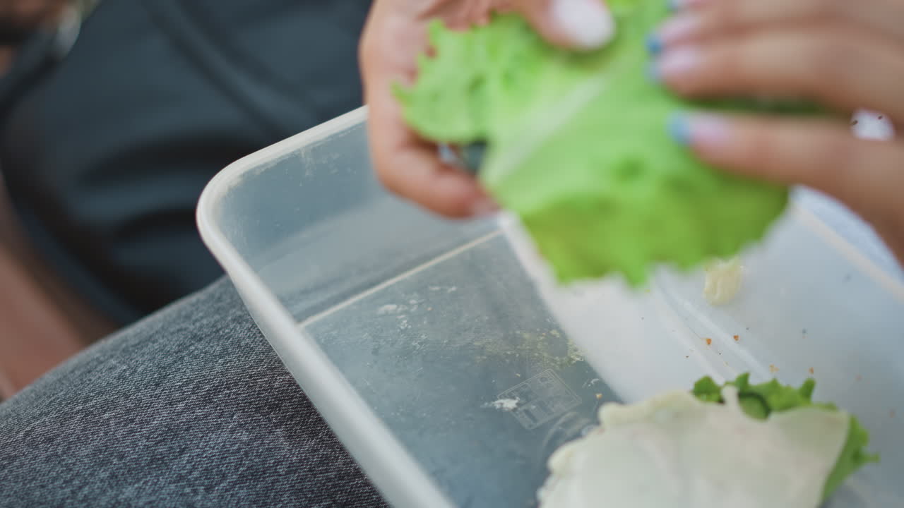 흰색 옷을 입은 여성 학생이 벤치에서 오렌지색 덮개를 들어 올려 신선한 양상추와 샌드위치를 보여 줍니다. 손질이 잘 된 손이 휴식 시간 동안 채소를 정리하는 모습이 클로즈업되어 있으며, 자연광과 캐주얼한 청바지가 편안한 분위기를 연출합니다.