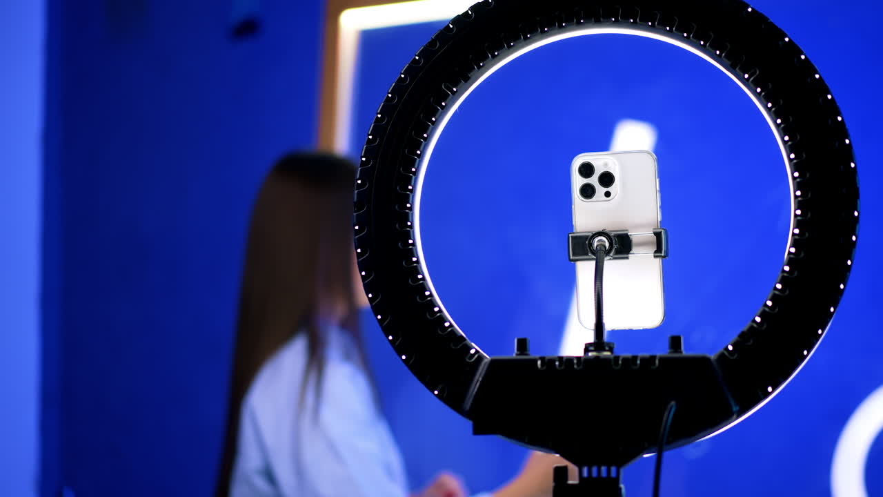 A smartphone set on tripod with a ring light above it. Long-haired lady stands at glass wall writing. Blurred backdrop.
