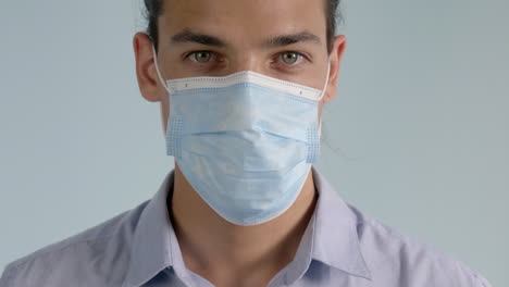 happy adult male with hazel eyes wearing collared button down shirt puts on surgical mask on a gray background to protect from covid-19 - coronavirus, straight on close up tripod shot