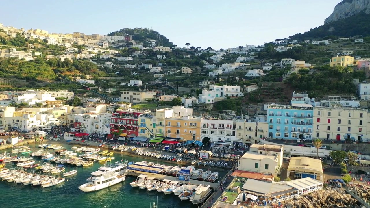 Ripresa aerea con drone di Marina Grande a Capri che mostra edifici colorati sul lungomare e un porto affollato di barche e case a terrazze che si ergono su un ripido pendio.