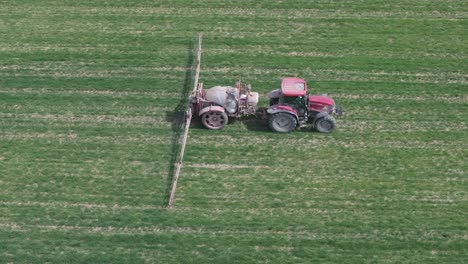 Vuelo-Lateral-Con-Dron-Filmando-Las-Labores-Agrícolas-Que-Realiza-Un-Agricultor-Con-Su-Tractor-Rojo-Y-Su-Remolque-Pulverizador-De-Grandes-Brazos-Extensibles-Sobre-Un-Campo-De-Cultivo-Con-Plantas-Verdes-Toledo