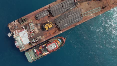 Aerial-flyover-industrial-crane-loading-pipes-on-old-rusted-ship-during-sunny-day-at-Gili-Trawangan-Pier