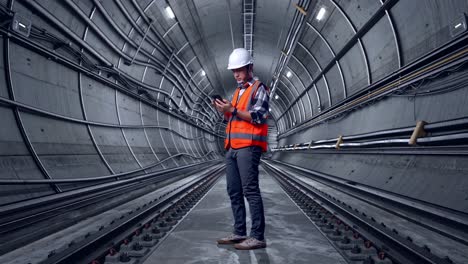 full body side view of asian male engineer with safety helmet using smartphone while standing in underground subway tunnel