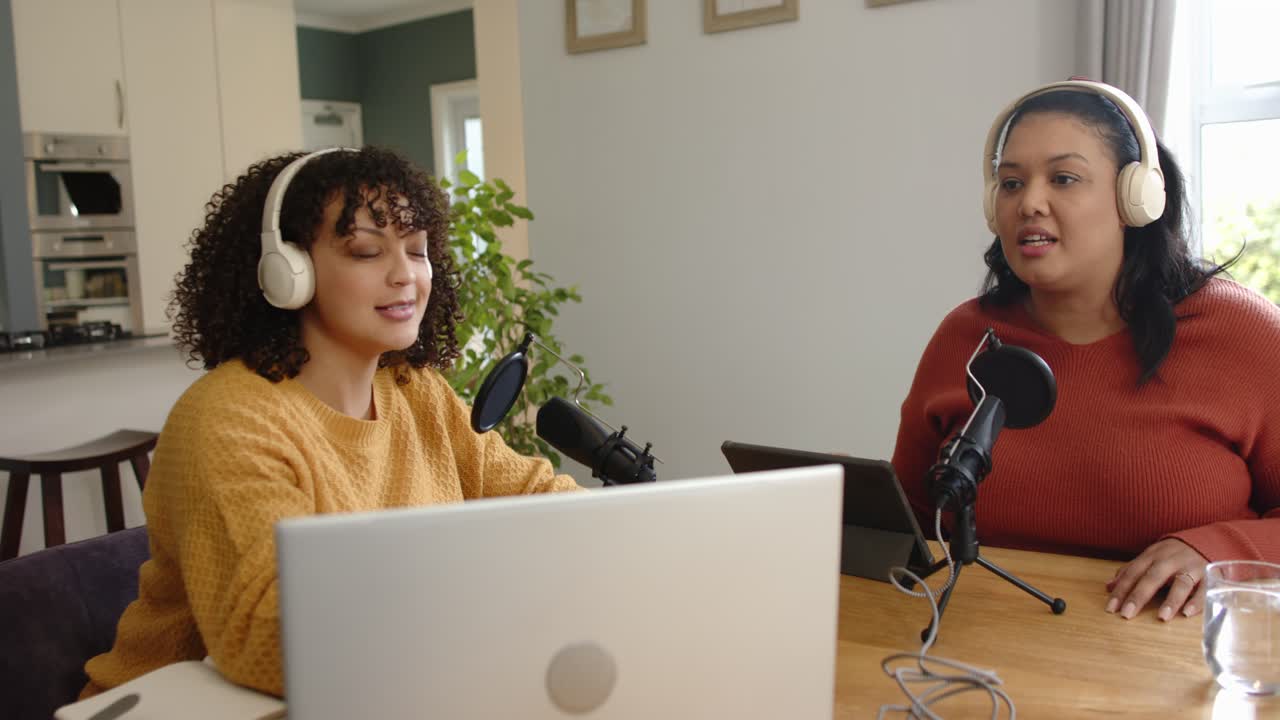Diverse female co-hosts testing audio and adjusting tablet while recording podcast in home kitchen