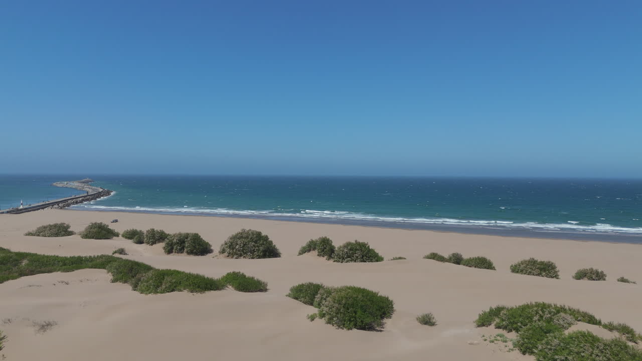 Forward motion drone shot over Necochea beach dunes, with plants in between and coastal barrier in background. Blue sea and sky. Shot on 4K-60fps.
