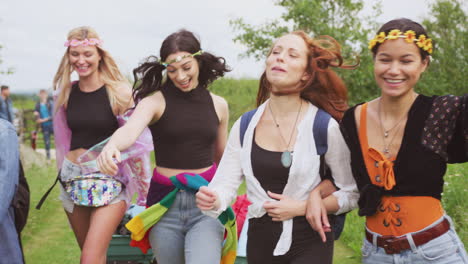 group of excited young female friends carrying camping equipment through field to music festival