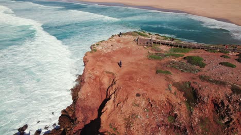 Flyover-view-of-female-hiker-revealing-the-ocean-waves-crashing-on-the-shoreline-of-Bordeira-surfing-beach-in-the-Algarve-region-of-Portugal-by-aerial-4k-drone