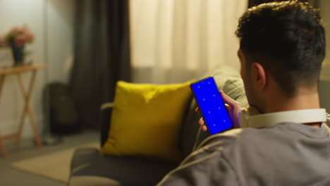 over the shoulder shot of man spending evening at home sitting on sofa with wireless headphones looking at blue screen mobile phone 1