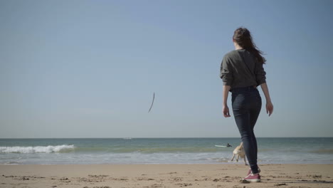 Young-woman-throwing-wooden-cane-to-dog-on-sandy-seashore.