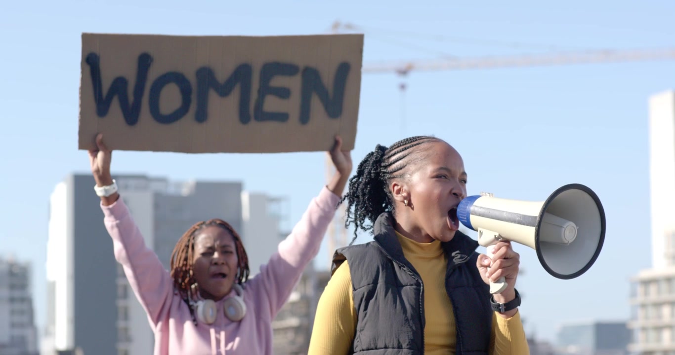 Eine afroamerikanische Frauenführerin erhebt die Faust, ruft in ein Megafon und hält ein „WOMEN“-Schild auf einem Dach.