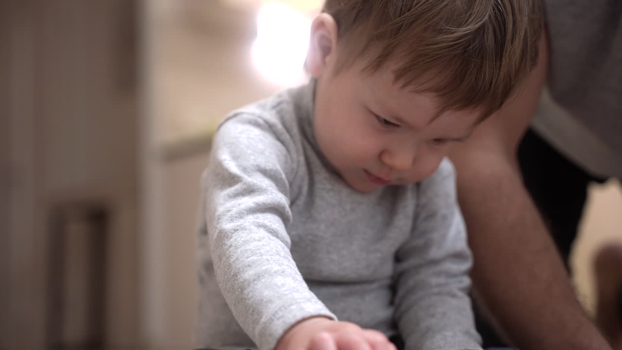 Mid shot of a baby sitting on the floor in the living room while his father playing with his toes
