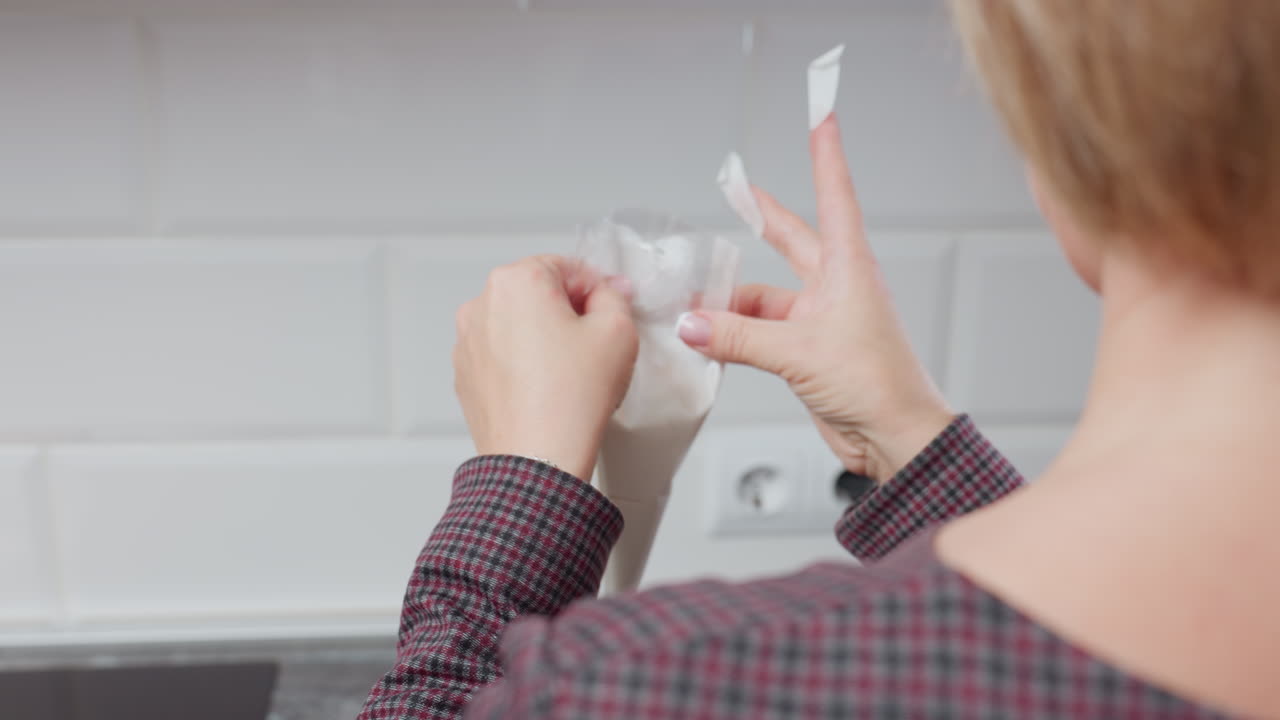 vista trasera de una mujer preparando algo en la cocina, sosteniendo una bolsa de tubería clara con crema o glaseado, centrada en las manos, la encimera y el fondo borroso