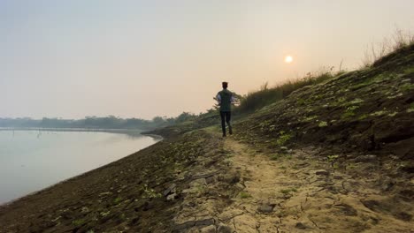 Vista-Trasera-Amplia-Del-Hombre-Trotando-En-El-Camino-Rural-Por-El-Lecho-Del-Río-En-Sylhet,-Mañana-Nebulosa