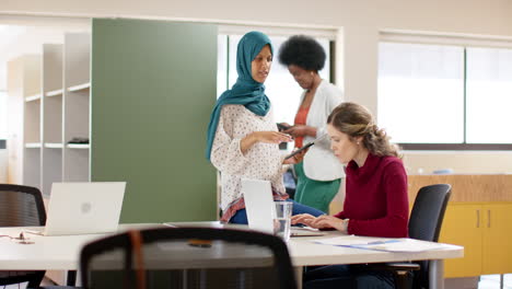 diverse female creative colleagues in discussion using laptop and tablet in office, slow motion