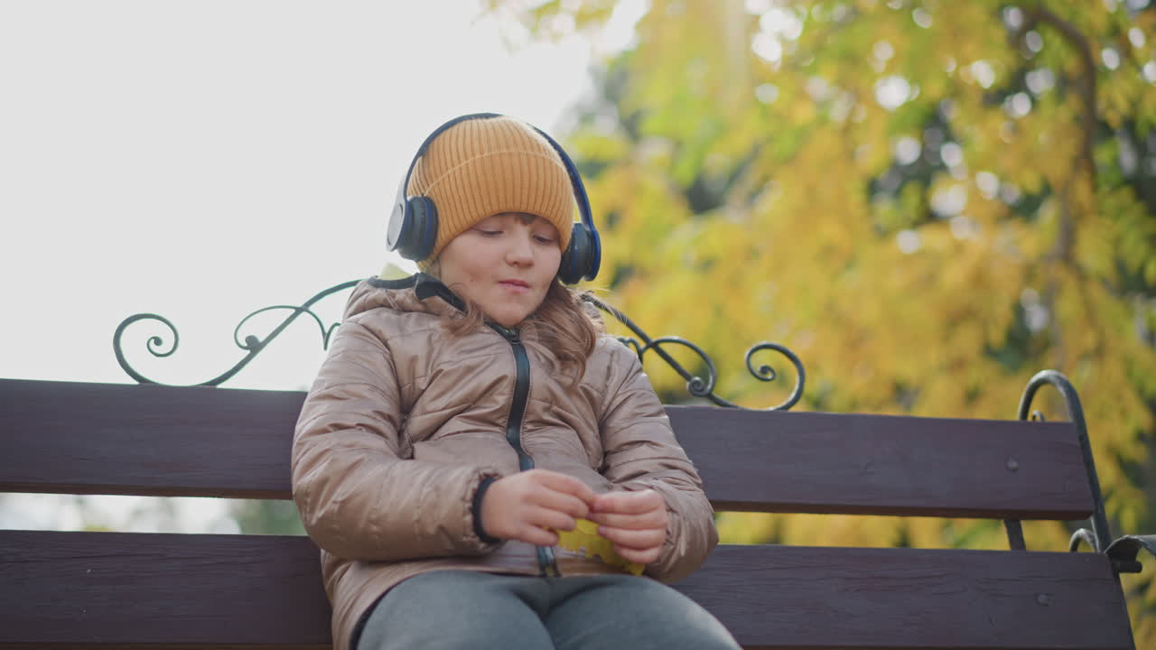 close up girl wearing beanie and headphones sitting on ornate bench in autumn park thoughtful gaze examining bright yellow leaves warm bokeh foliage backdrop scattered leaves