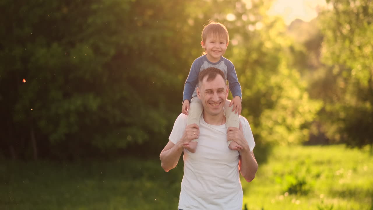 padre amoroso sonríe caminando con el niño sentado en el cuello al atardecer en un prado en verano en cámara lenta.